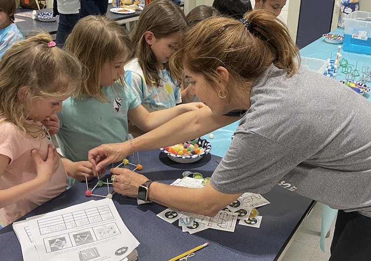 Seorang wanita bersandar di seberang meja untuk membantu salah satu dari beberapa anak kecil membangun sebuah struktur kubus dari bola-bola kecil berwarna-warni dan tongkat-tongkat.